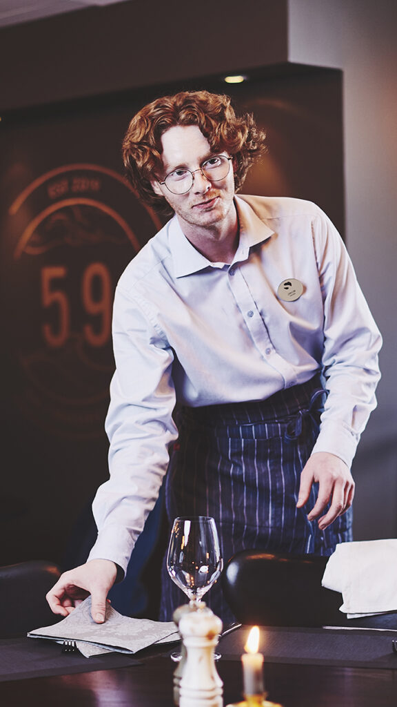 Servitør med raudt krøllete hår legg ein serviett på bordet og smiler lurt.